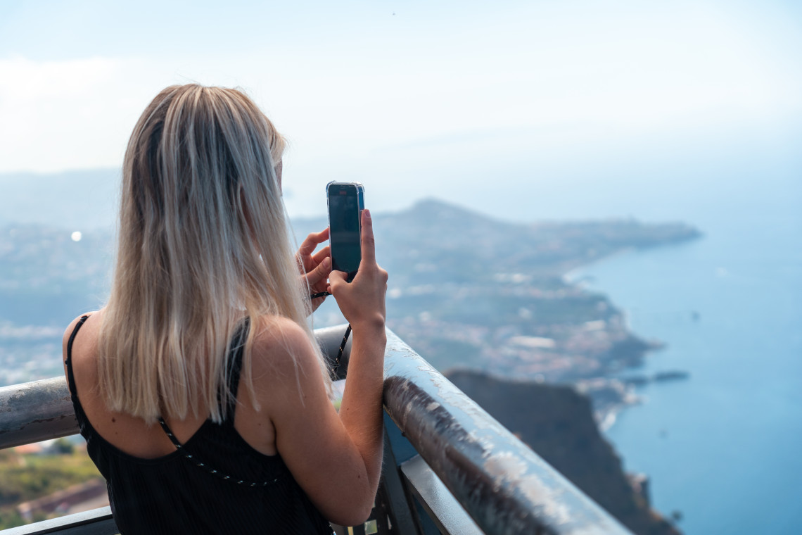 Visitor photographing the mountainous landscape and Atlantic coastline from an elevated viewpoint on Madeira Island.