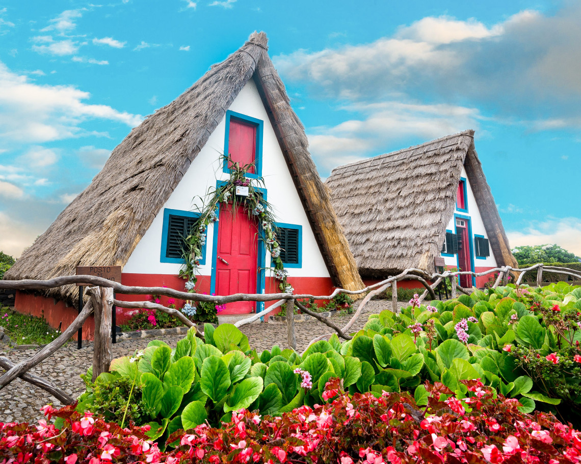 Traditional thatched houses in Santana, Madeira, Portugal, feature steep triangular roofs, white walls, red doors, and blue window frames. Colorful flower beds and rustic wooden fences surround these distinctive examples of Madeira&rsquo;s rural architecture.