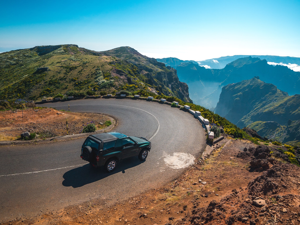 A four-wheel-drive vehicle follows a sharp mountain road near Pico do Arieiro in Madeira, Portugal. The route overlooks deep volcanic valleys, rugged ridges, and low clouds across the island&rsquo;s central highlands.