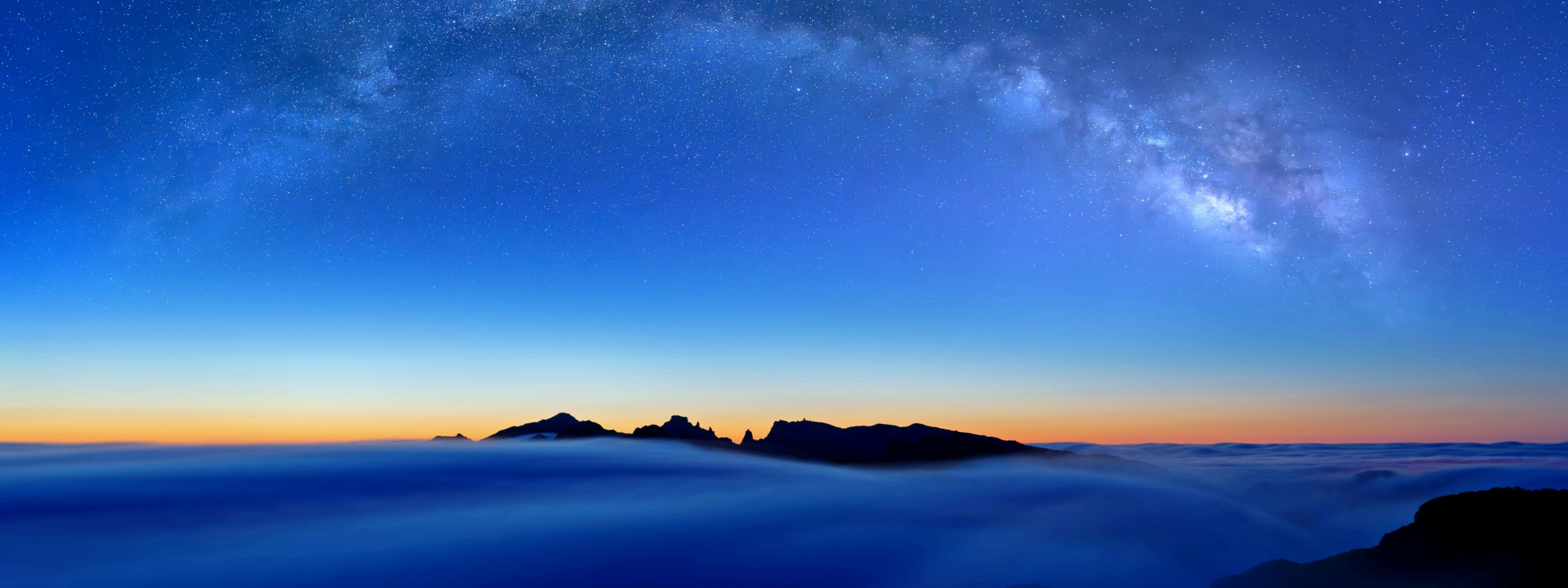 The Milky Way stretches across the night sky above Pico do Arieiro on Madeira Island, Portugal. Dark mountain peaks rise above a dense layer of clouds, with a narrow band of sunset light visible on the horizon.