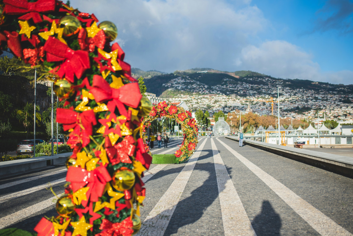 Red and gold Christmas decorations line the waterfront promenade in Funchal, Madeira Island, Portugal. The decorated walkway leads toward the city&rsquo;s hillside neighbourhoods beneath a partly cloudy sky.