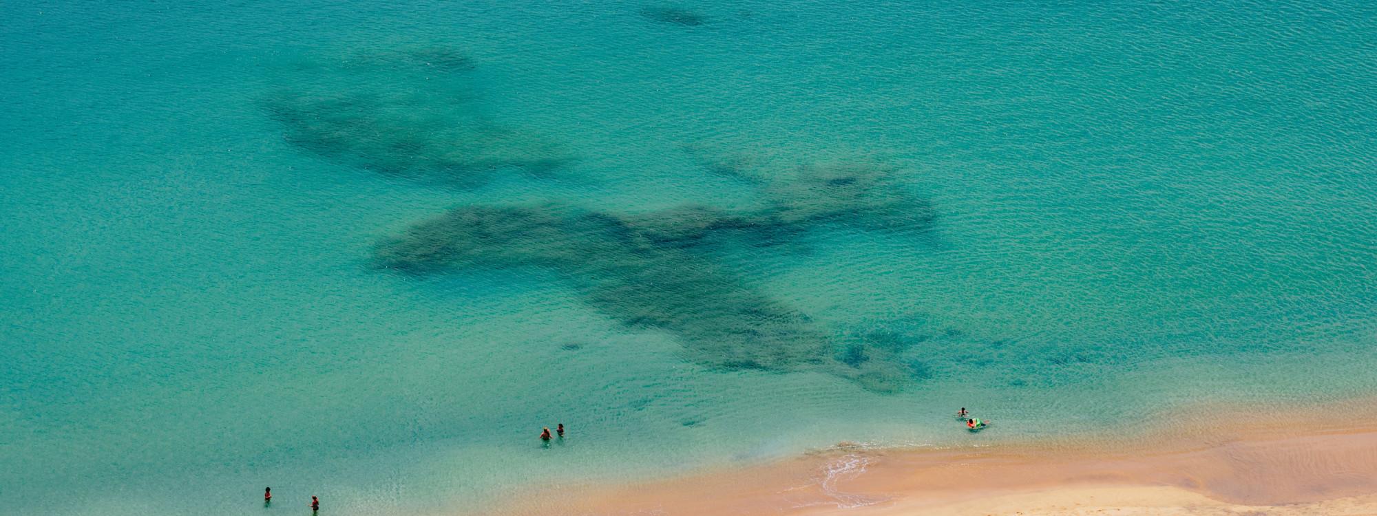 Aerial view of swimmers in the clear turquoise water at Porto Santo Beach near Vila Baleira, Portugal. Dark underwater rock formations contrast with the island’s pale golden sand along the Atlantic shoreline.
