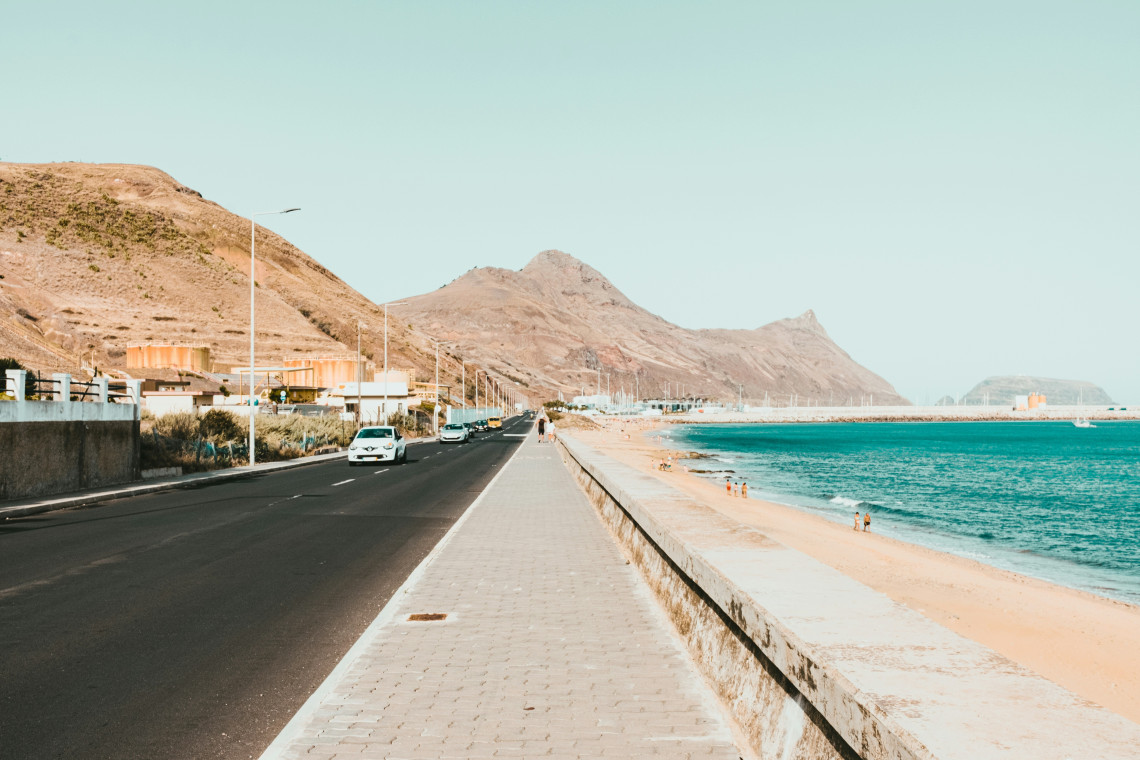 A coastal road and pedestrian promenade run beside Porto Santo Beach in Vila Baleira, Portugal, with golden sand and turquoise Atlantic water on one side. The dry mountain landscape of Porto Santo rises behind the town and shoreline.