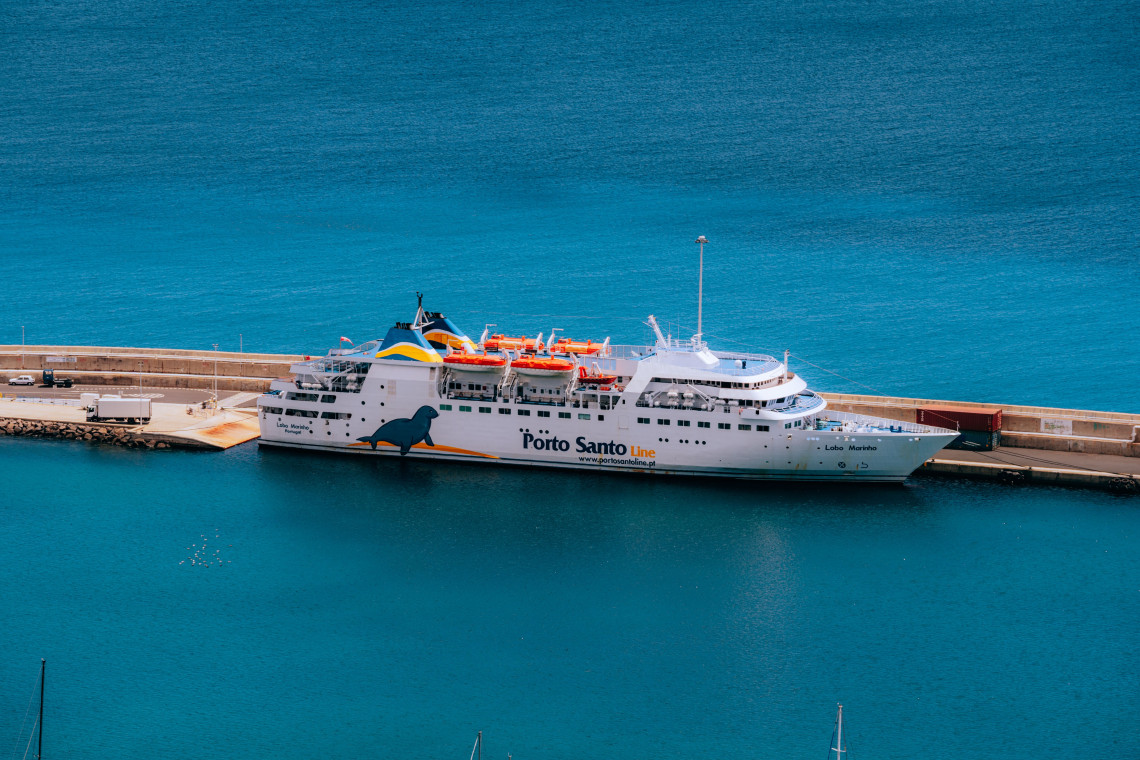 The Porto Santo Line ferry is docked at the port of Vila Baleira on Porto Santo Island, Portugal. The passenger vessel operates across the Atlantic between Madeira and Porto Santo, with orange lifeboats visible along its upper deck.