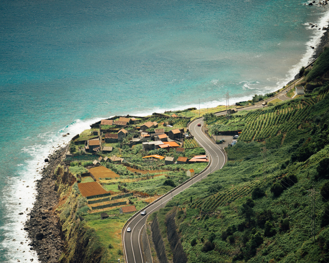 Elevated view of Faj&atilde; dos Padres in C&acirc;mara de Lobos, Madeira, Portugal, showing terraced farmland, traditional houses, and a winding coastal road beside the Atlantic Ocean. The cultivated strip lies at the base of Madeira&rsquo;s steep southern cliffs.
