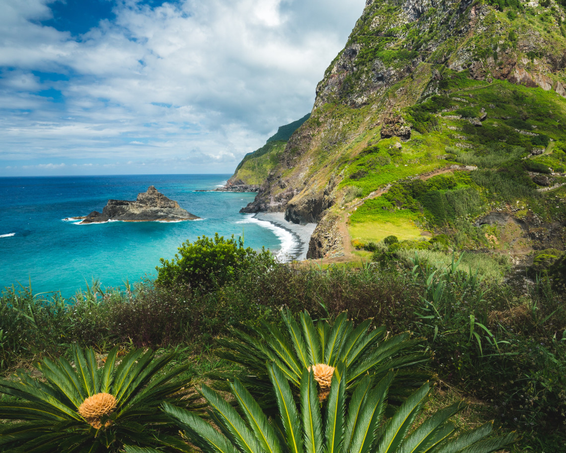 A winding trail crosses the steep green cliffs of Boaventura on Madeira&rsquo;s northern coast, with a rocky islet rising from the Atlantic Ocean below. Subtropical vegetation frames the view of the secluded shoreline and rugged volcanic landscape.