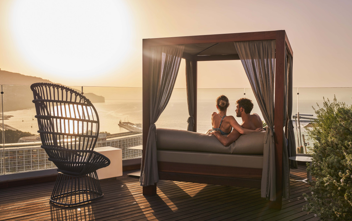 Guests relax on a terrace at Savoy Palace in Funchal, Madeira Island, Portugal, overlooking the Atlantic Ocean and the city coastline at sunset. The image shows a luxury hotel experience with panoramic sea views in Madeira&rsquo;s capital.