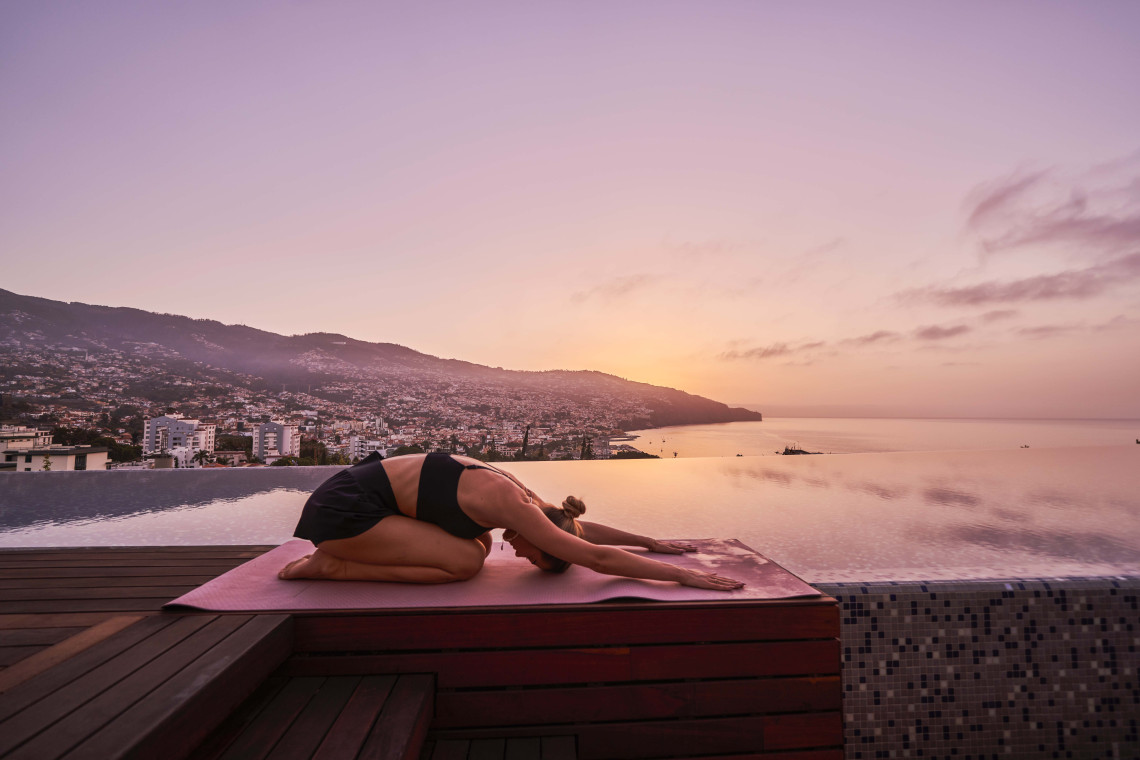 A traveler practices yoga beside an infinity pool in Funchal, Madeira, Portugal, overlooking the Atlantic Ocean at sunset. The image highlights a peaceful wellness experience with views over the city and coastline.
