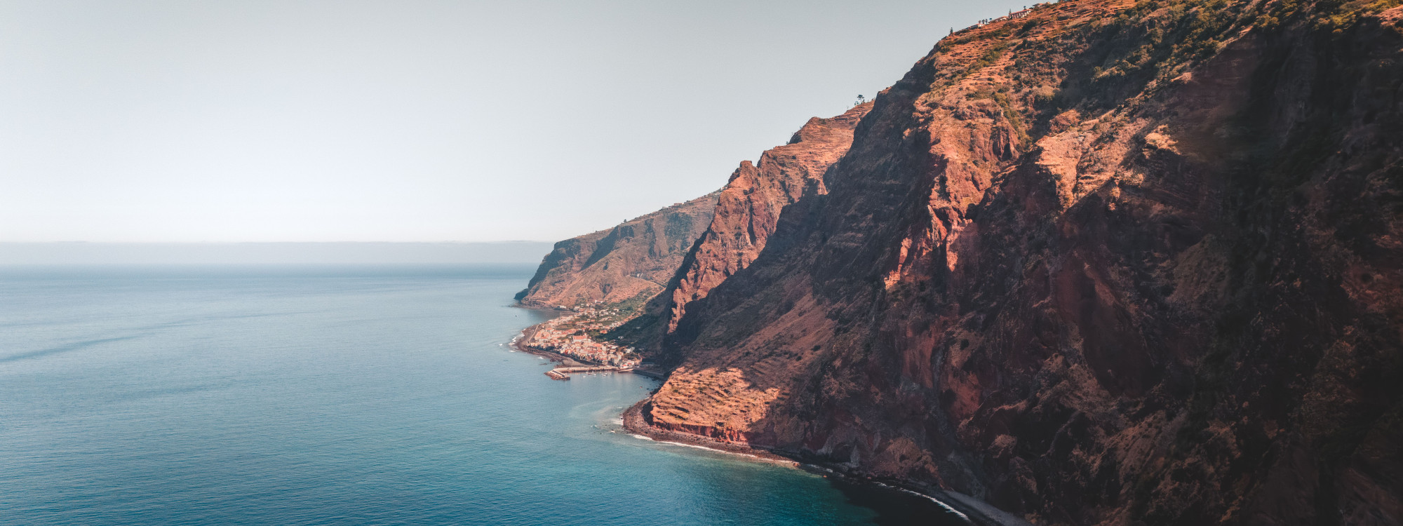 View along the rugged coastline near Cabo Girão in Câmara de Lobos, Madeira, Portugal, showing steep reddish volcanic cliffs descending into the Atlantic Ocean. A narrow coastal road follows the base of the cliffs beside the calm blue water.