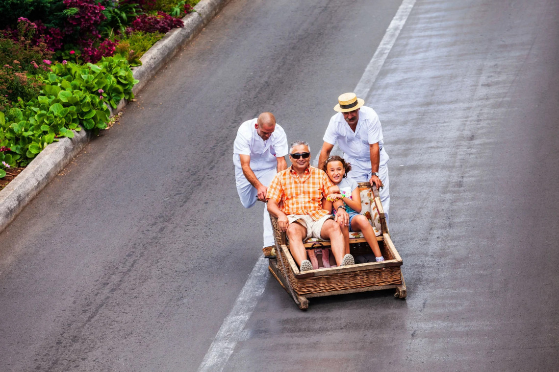 Two carreiros guide a father and daughter downhill in a traditional wicker toboggan in Monte, Funchal, Madeira, Portugal. The image shows the historic street ride and the wooden runners used to steer the basket