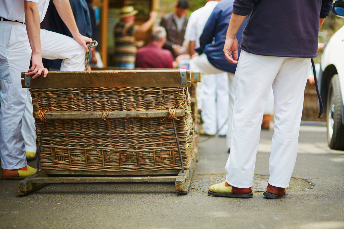 Monte carreiros stand beside a traditional wicker toboggan in Funchal, Madeira, Portugal. The woven basket, wooden runners, and distinctive white uniforms reflect the island&rsquo;s historic downhill sledge tradition.