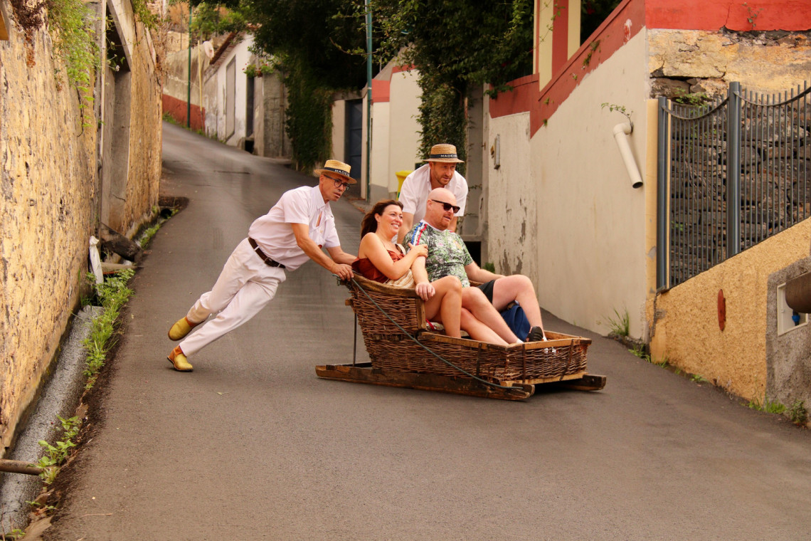 Two carreiros guide visitors downhill in a traditional wicker toboggan through the narrow streets of Monte in Funchal, Madeira, Portugal. The image shows the historic basket sled, wooden runners, and distinctive white uniforms used during the descent.