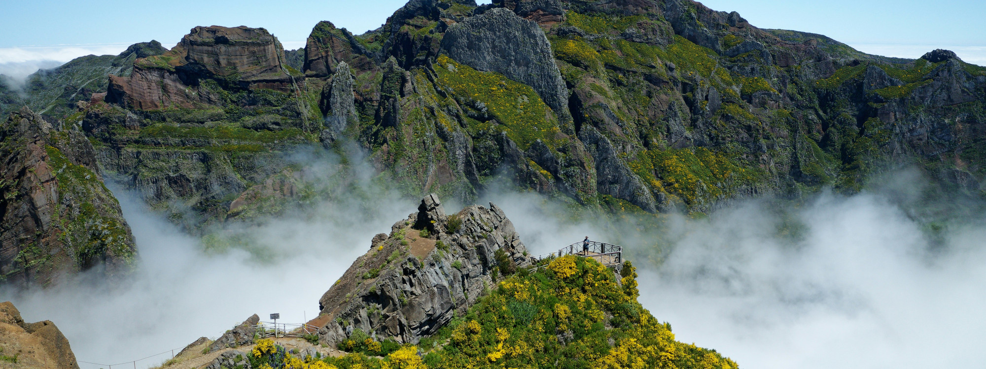 Rocky mountain peaks rise through low clouds at Pico do Arieiro in Madeira, Portugal. The view shows the island’s rugged central mountain range, with steep ridges covered in green and yellow vegetation.