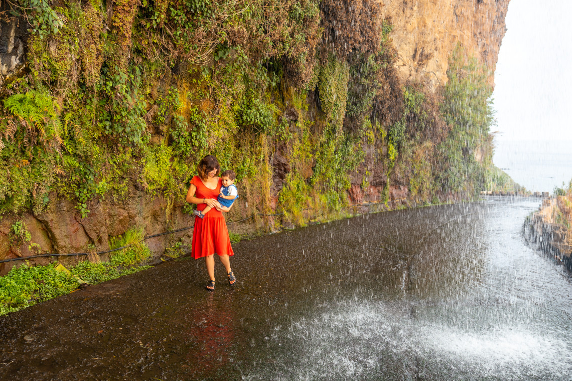 A woman holding a child stands beneath Cascata dos Anjos in Ponta do Sol, Madeira, Portugal. The waterfall flows directly over a coastal road beside a steep, vegetation-covered cliff.