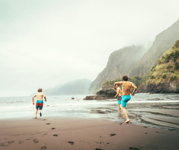 Two people run along the black sand of Seixal Beach in Madeira, Portugal, with steep volcanic cliffs and the Atlantic Ocean in the background. The scene highlights one of the island’s most distinctive natural beaches and its dramatic coastal landscape.