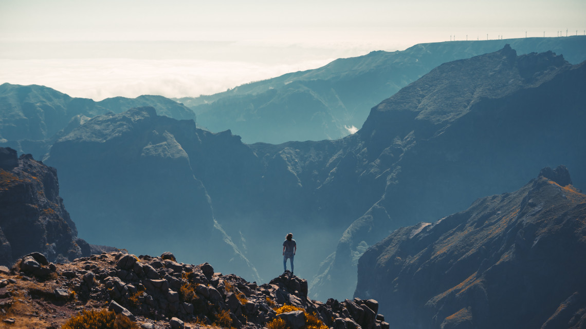 A hiker stands on a rocky viewpoint at Pico do Arieiro in Madeira, Portugal, overlooking layered volcanic peaks and deep mountain valleys. The elevated landscape reveals the rugged terrain of Madeira&rsquo;s central mountain range beneath clear daylight.