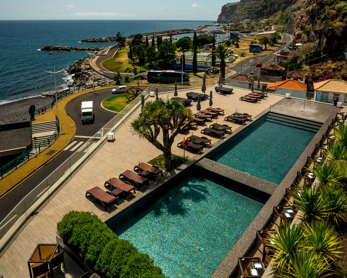 Elevated view of the outdoor swimming pools at Saccharum Resort in Calheta, Madeira, Portugal. Sun loungers overlook the Atlantic coast, the seafront promenade, and Calheta Marina beneath steep volcanic cliffs.