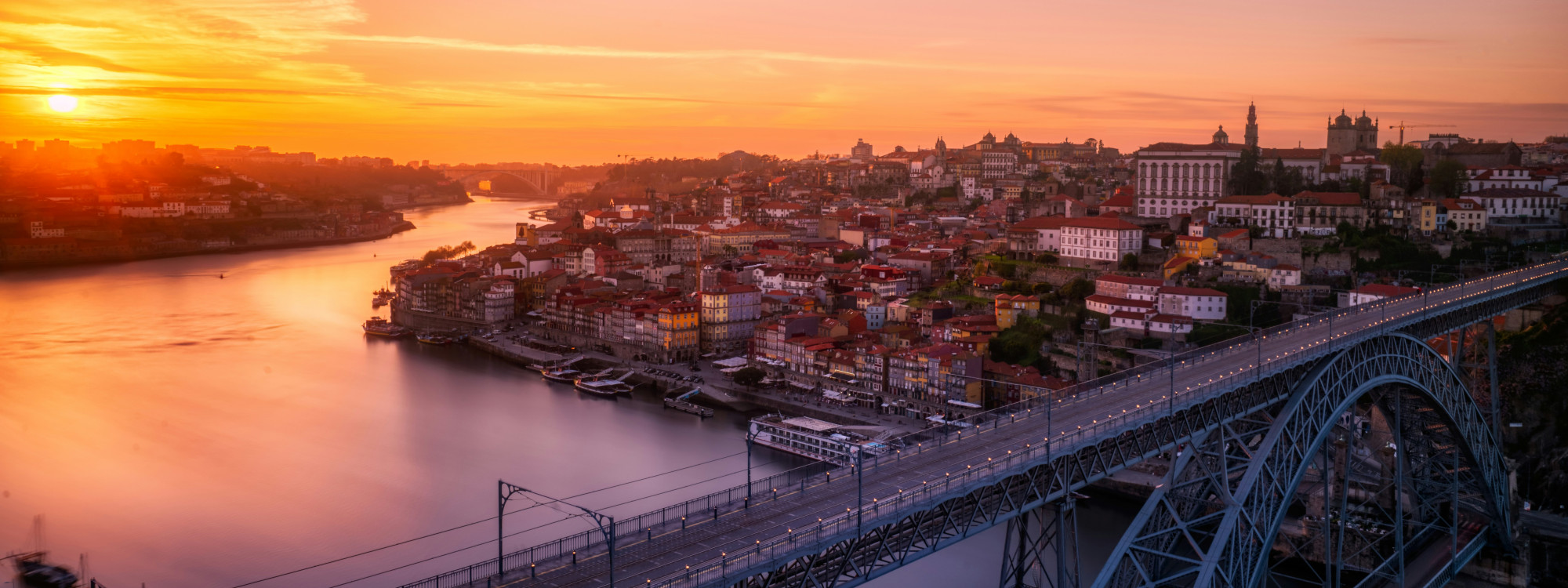 Sunset view of Dom Luís I Bridge in Porto, Portugal, crossing the Douro River beside the historic Ribeira waterfront. The image shows Porto’s riverside architecture and warm evening light over the city.