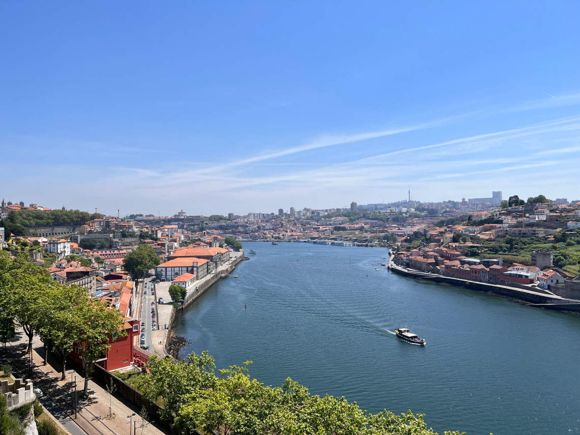 Panoramic view of the Douro River flowing between Porto and Vila Nova de Gaia, Portugal. A sightseeing boat crosses the water between terracotta-roofed neighbourhoods and historic riverside buildings.