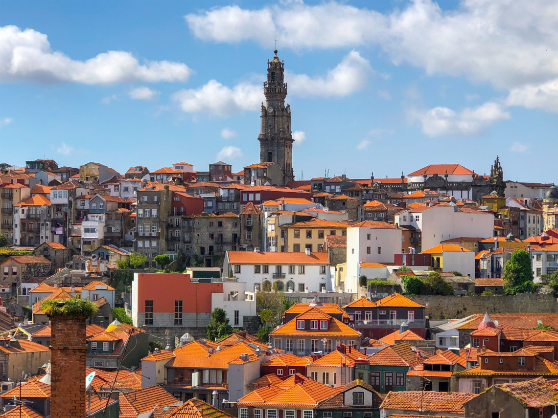 Cl&eacute;rigos Tower rises above the terracotta rooftops and traditional buildings of central Porto, Portugal. The Baroque bell tower stands against a blue sky with scattered white clouds.