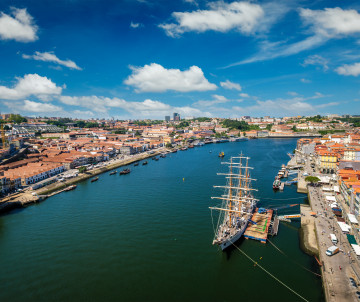Aerial view of the Douro River between Porto and Vila Nova de Gaia, Portugal, with boats travelling through the waterway. Terracotta-roofed neighbourhoods, riverside promenades and a tall sailing ship line both banks.