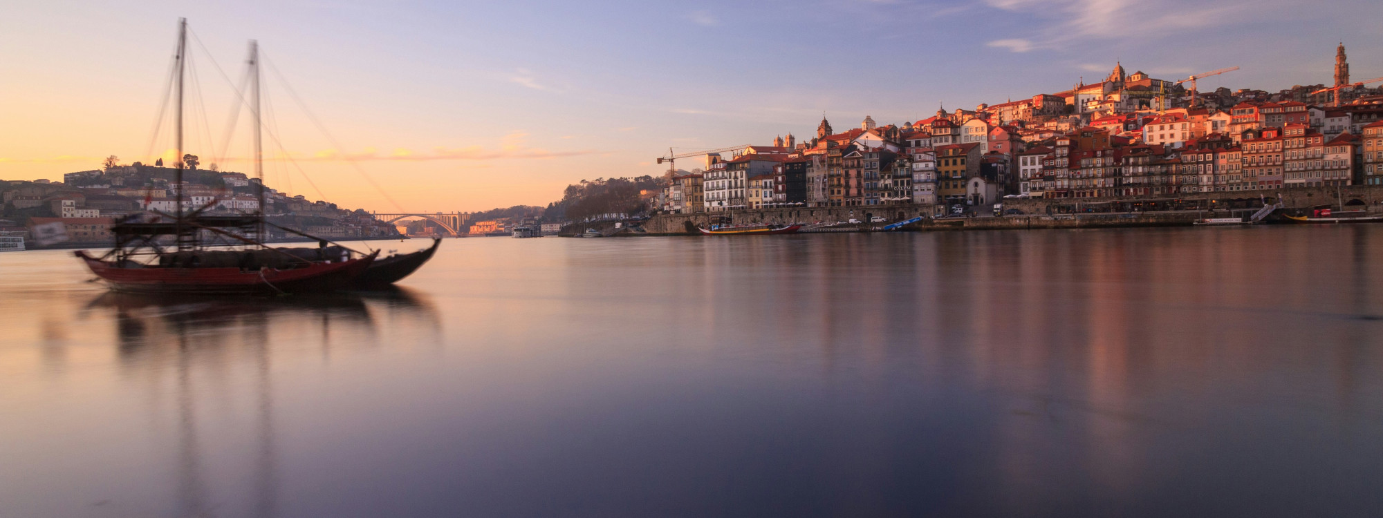 Sunset view of the Ribeira waterfront in Porto, Portugal, along the Douro River. A traditional rabelo boat is seen on the water with Porto’s historic riverside buildings in the background.