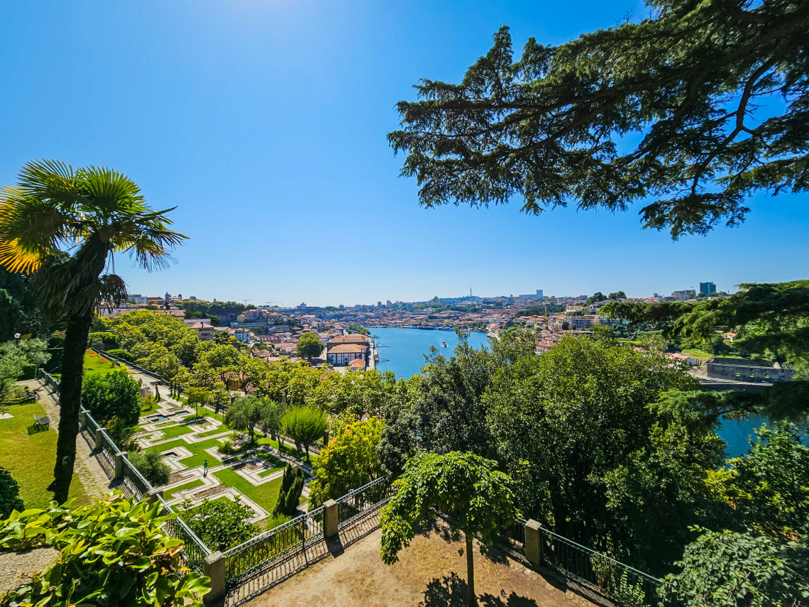 View from Jardins do Pal&aacute;cio de Cristal in Porto, Portugal, overlooking the Douro River, city rooftops, and surrounding gardens. The image shows one of Porto&rsquo;s scenic viewpoints with greenery framing the river landscape.