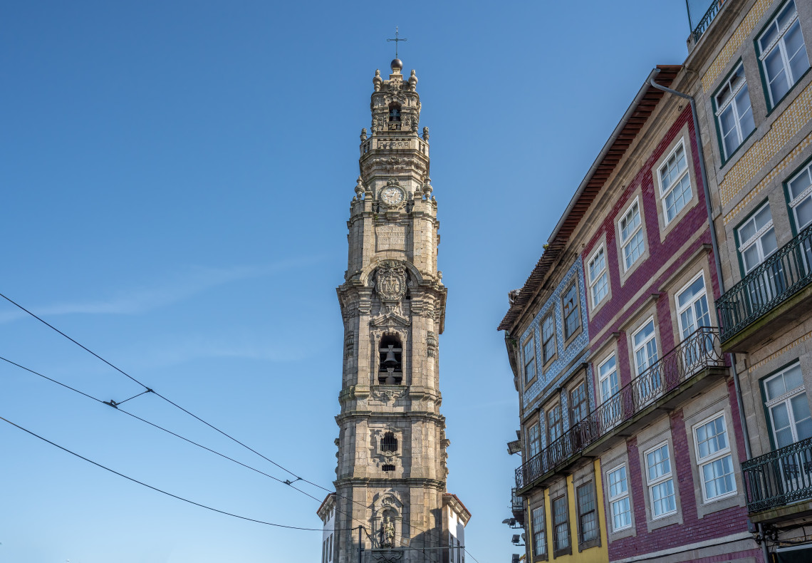 View of Cl&eacute;rigos Tower in Porto, Portugal, rising above traditional city buildings. The image shows the baroque bell tower against a clear blue sky in Porto&rsquo;s historic center.