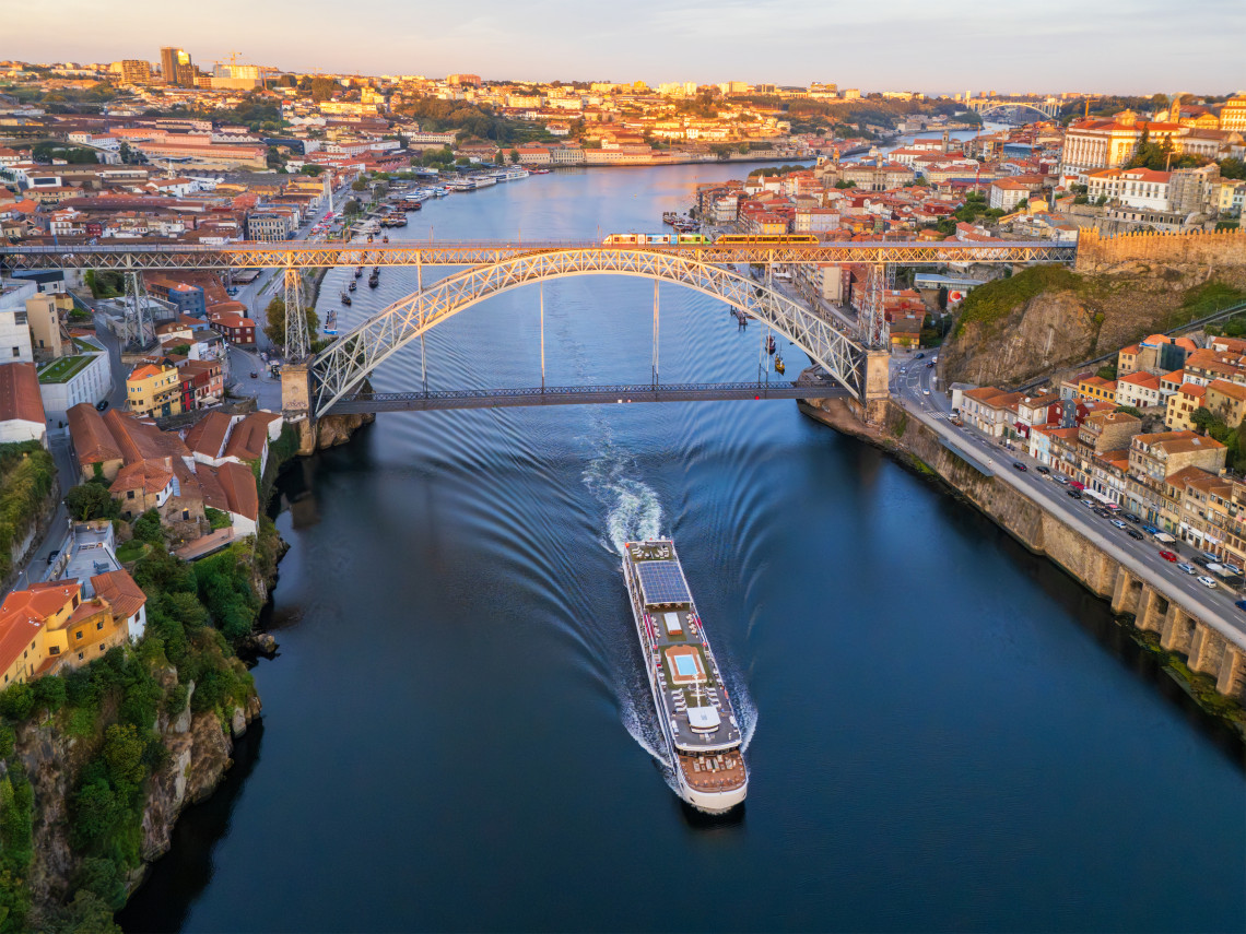 Aerial view of Dom Lu&iacute;s I Bridge in Porto, Portugal, spanning the Douro River between Porto and Vila Nova de Gaia. A river cruise boat passes beneath the iron bridge with the city&rsquo;s historic riverside districts on both banks.