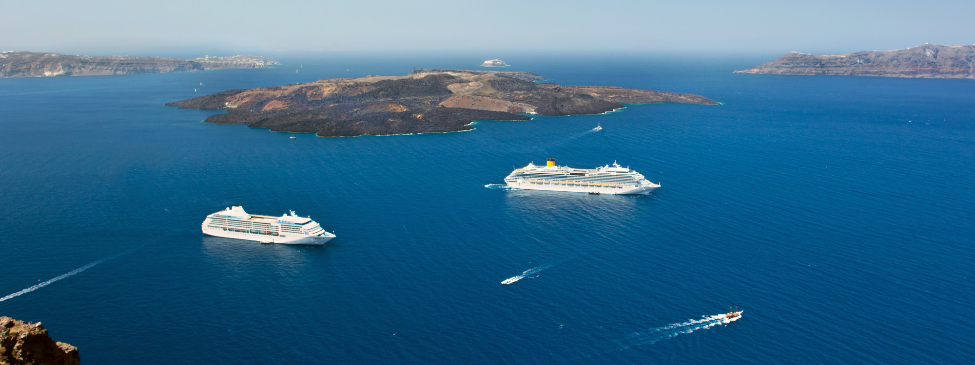 Multiple cruise ships sailing through deep blue waters between rocky Mediterranean islands in Greece, viewed from an elevated coastal viewpoint.