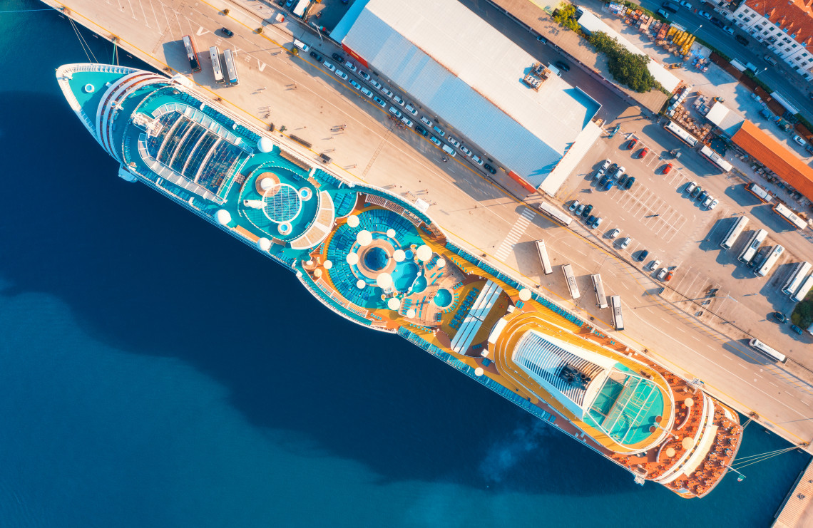 Top-down aerial view of a large cruise ship docked at the Port of Barcelona, Spain, showing the vessel&rsquo;s open decks, pools and terminal facilities beside the Mediterranean Sea.