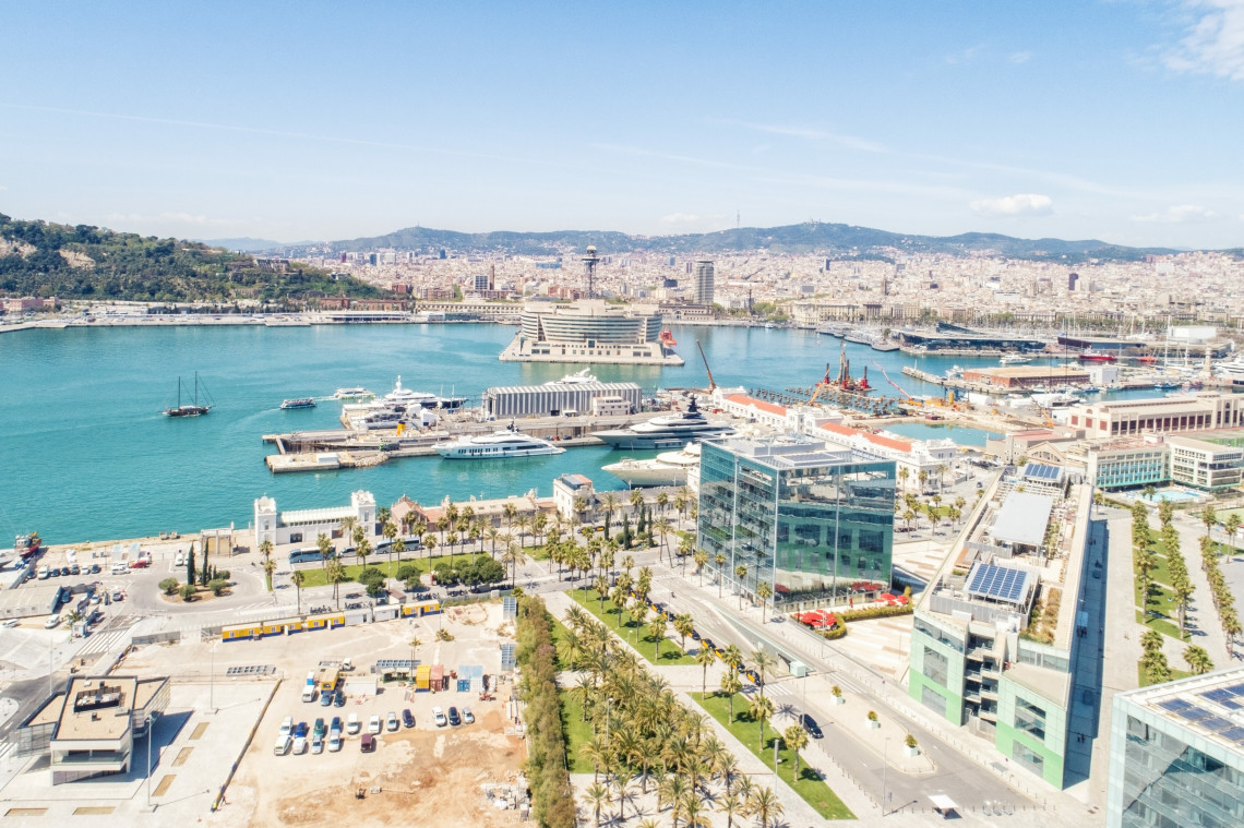 Elevated view across Port Vell in Barcelona, Spain, with marina waters, passenger terminals, waterfront buildings and the city skyline surrounding the harbour.