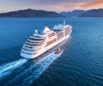 Large cruise ship sailing across calm Mediterranean waters at sunset, with mountainous coastline visible in the distance.
