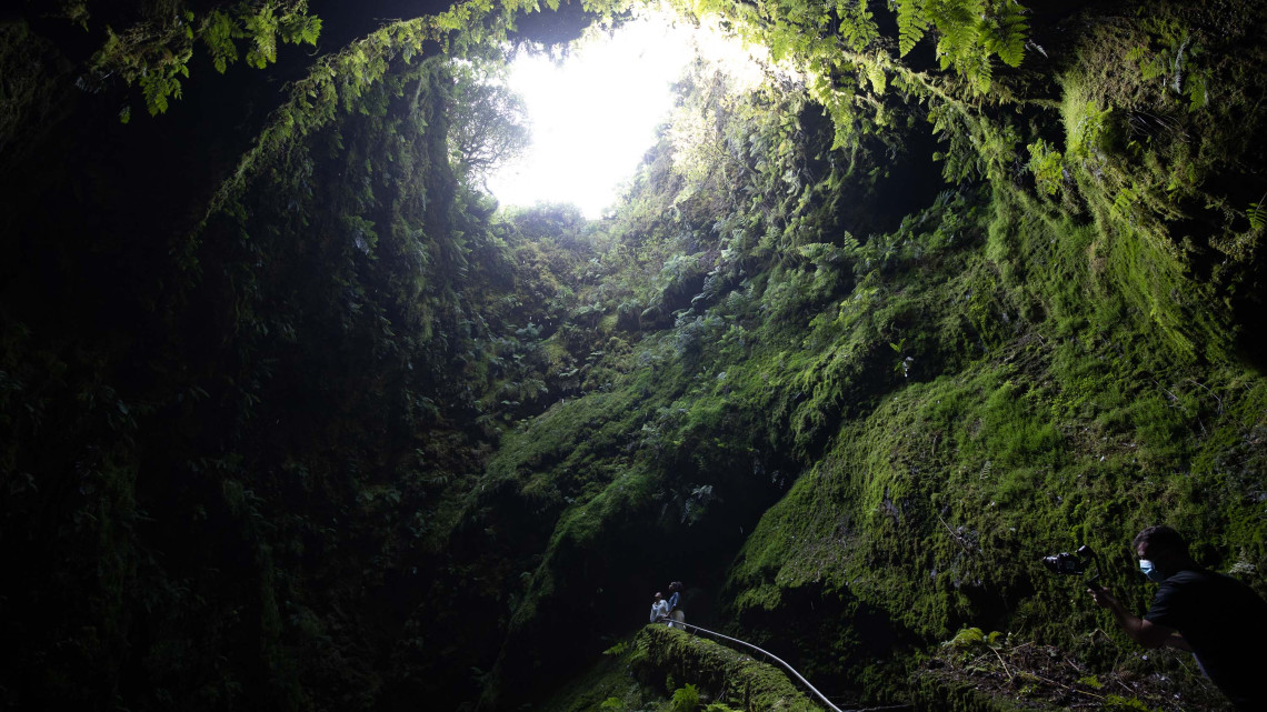 Visitors standing inside the Algar do Carv&atilde;o lava tube looking up through the volcanic chimney opening surrounded by moss-covered walls, Terceira Island, Azores