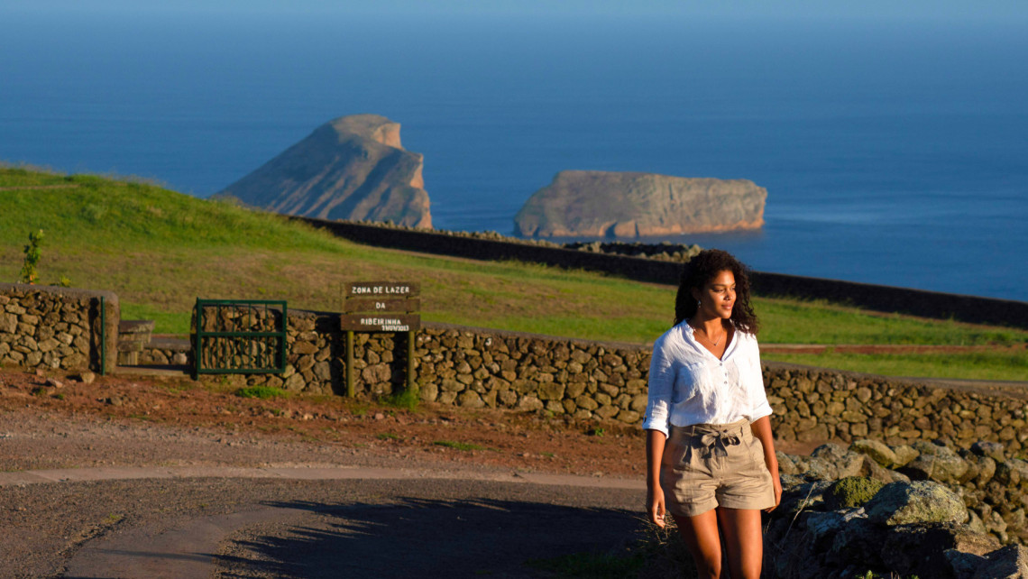 Woman walking along a scenic coastal road in Ribeirinha with traditional basalt stone walls, green hillside, and volcanic islets rising from the Atlantic Ocean, Terceira Island, Azores