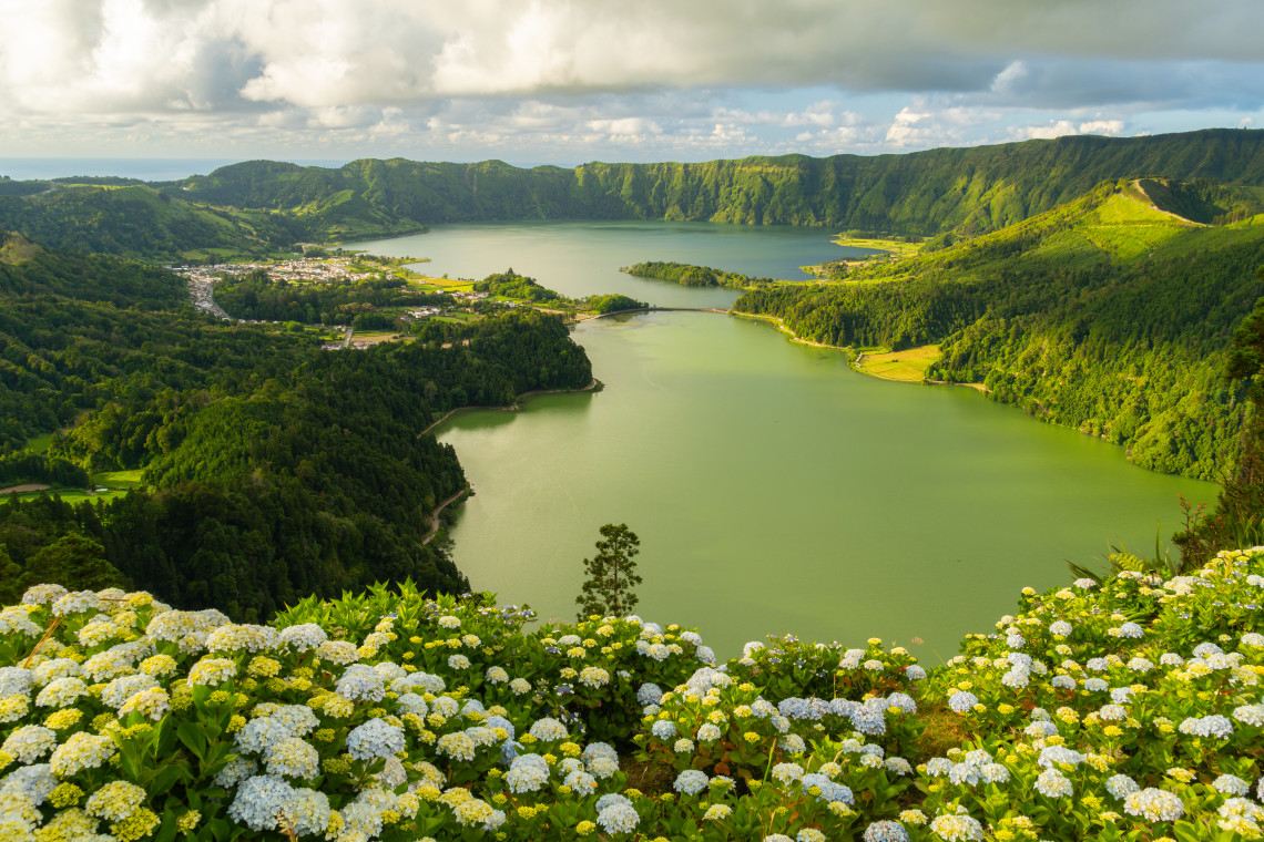 View from Vista do Rei viewpoint over the Sete Cidades caldera on S&atilde;o Miguel Island, Azores, with white hydrangeas in the foreground, Lagoa Verde's green water below, the village of Sete Cidades on the left, and Lagoa Azul stretching into the distance under a dramatic cloudy sky
