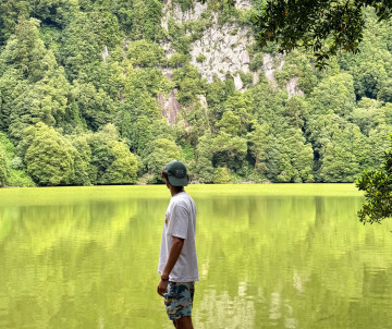 A man standing at the edge of Lagoa do Congro on São Miguel Island, Azores, looking across the vivid green water toward steep forested volcanic cliffs rising from the shoreline