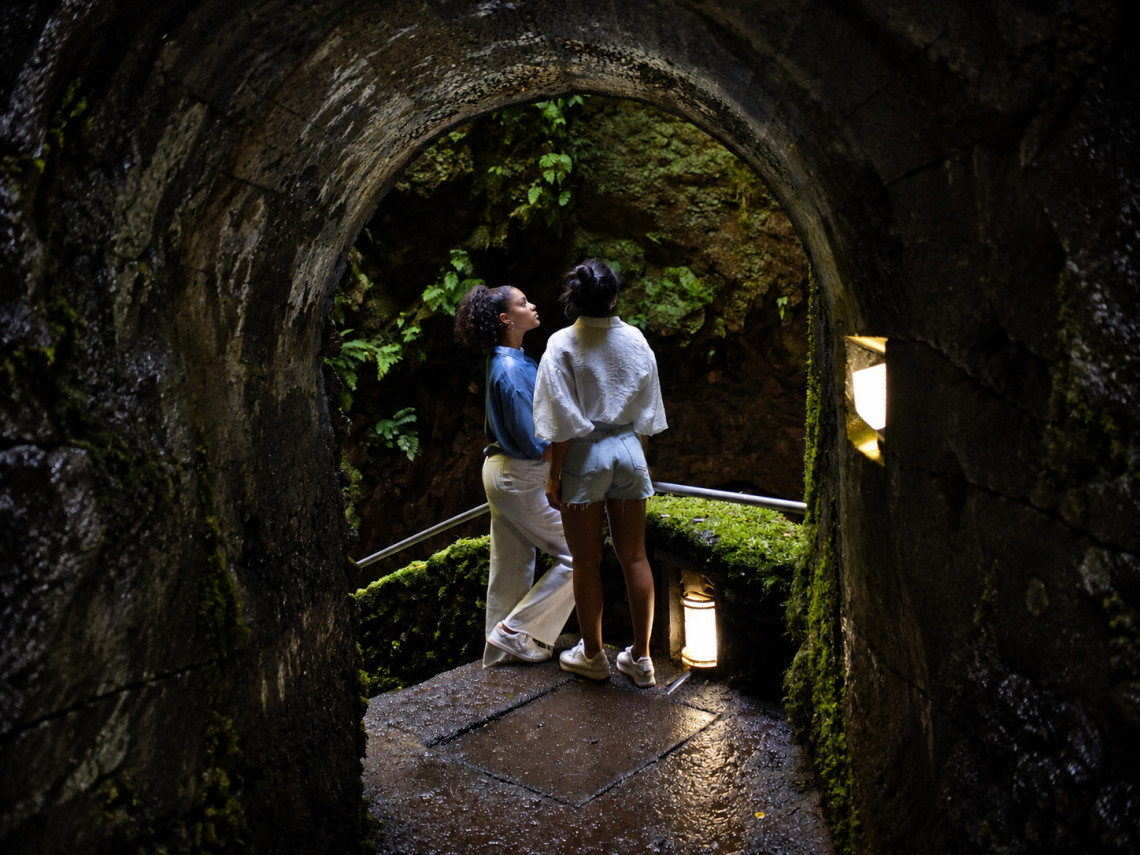 Visitors exploring the underground walking route inside Algar do Carv&atilde;o on Terceira Island, framed by a dark stone passage covered with moss and ferns.