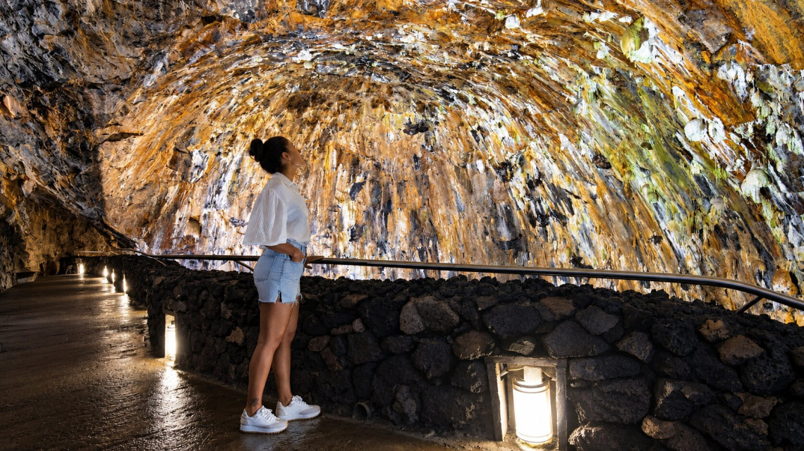 Visitor observing the colorful mineral-covered volcanic walls inside Algar do Carv&atilde;o on Terceira Island, illuminated along the underground walking path.