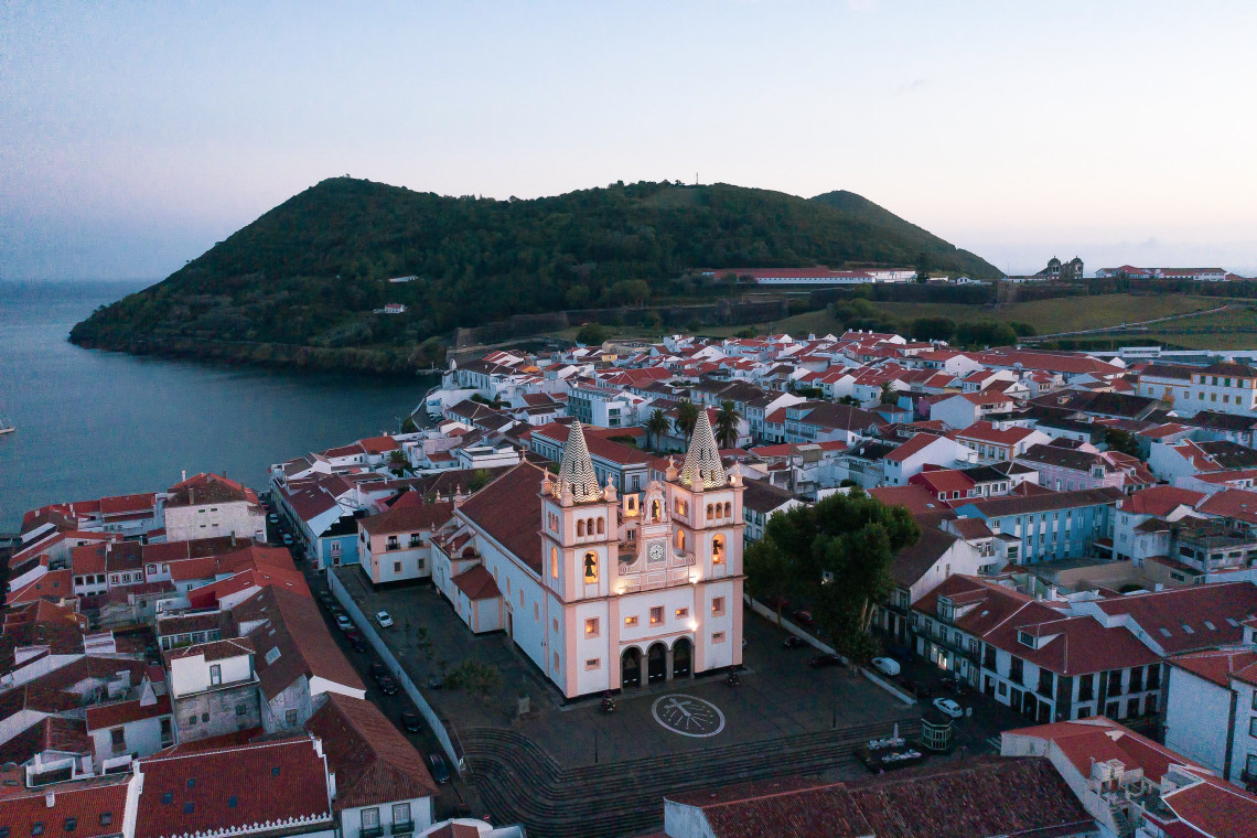 Aerial view of S&eacute; Catedral de Angra do Hero&iacute;smo rising above the red-tiled rooftops of the historic city center, with its twin patterned spires and broad stone plaza.