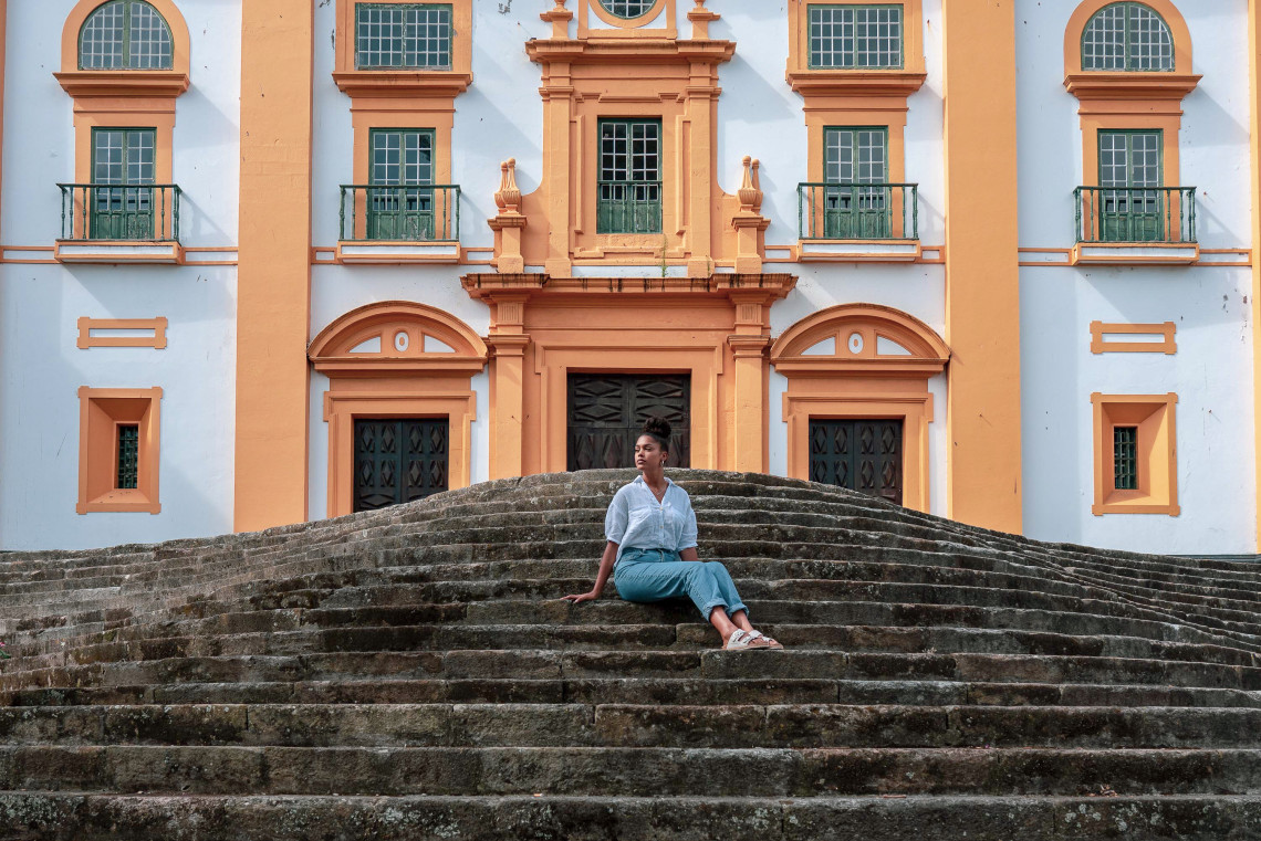 Traveler sitting on the stone steps of Igreja de Nossa Senhora do Carmo in Angra do Hero&iacute;smo, framed by the church&rsquo;s symmetrical white-and-ochre fa&ccedil;ade and tall windows.