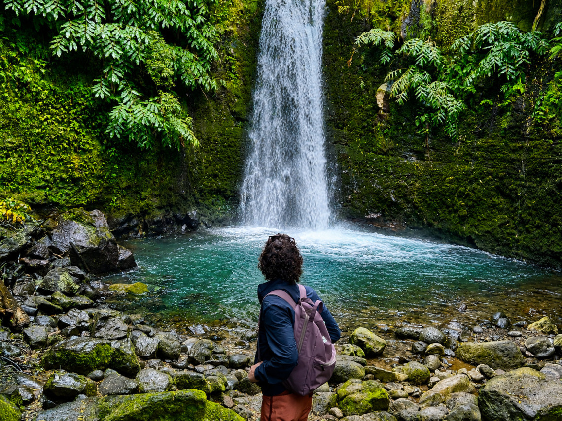 A traveler with a backpack stands on a rocky shore facing Salto do Prego Waterfall in Sanguinho, S&atilde;o Miguel Island, Azores, surrounded by lush green vegetation and a clear pool.