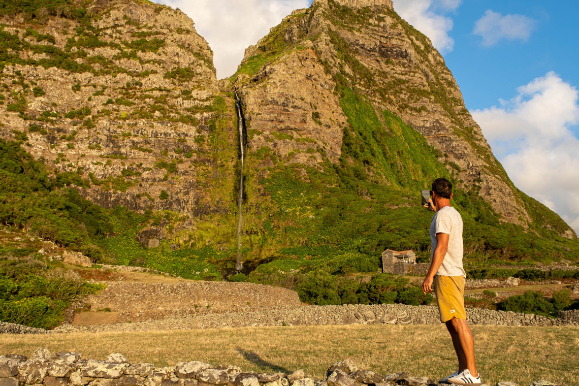 Visitor photographing Cascata do Po&ccedil;o do Bacalhau on Flores Island, Azores, Portugal. The tall waterfall descends from steep volcanic cliffs into a lush valley surrounded by native vegetation.