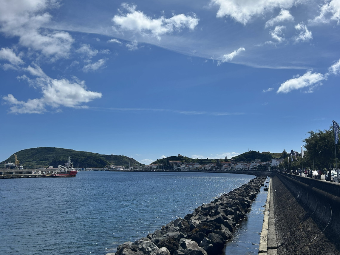 View of Horta on Faial Island, Azores, Portugal, with the waterfront promenade, marina, and harbour along the Atlantic coast. The town stretches across the bay beneath green volcanic hills and a clear blue sky.