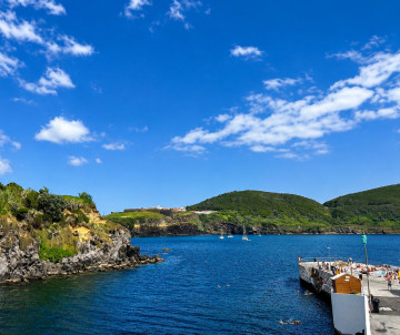 Natural swimming pools at Piscinas da Silveira in Terceira Island, Azores, with blue ocean water, rocky coastline, green hills, and a sunny sky.