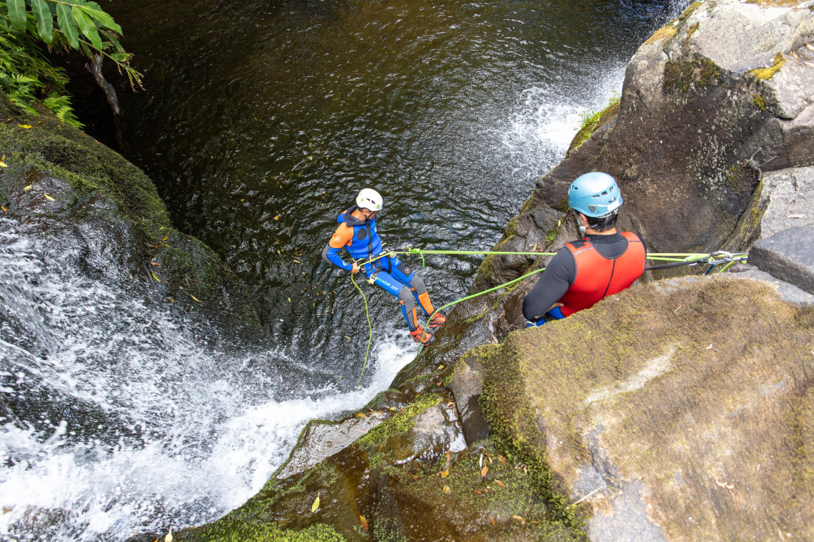 A canyoning participant rappels down a waterfall on Flores Island while a guide manages the ropes from the volcanic rocks above.
