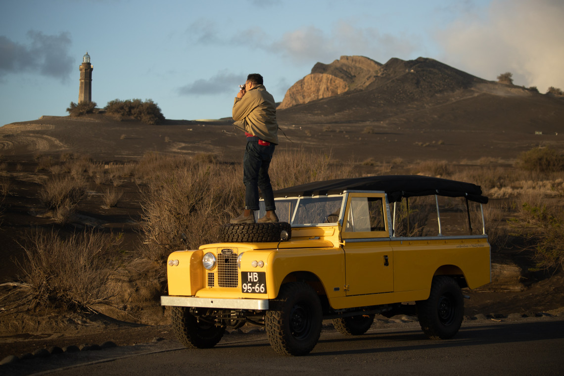 A traveler stands on a yellow off-road vehicle beside the volcanic landscape of Capelinhos on Faial Island, with the historic lighthouse and dark eruption terrain in the background.