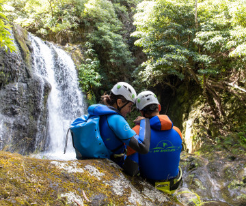 Two canyoning participants wearing helmets and wetsuits rest beside a waterfall on Flores Island, surrounded by dense green vegetation and volcanic rock.