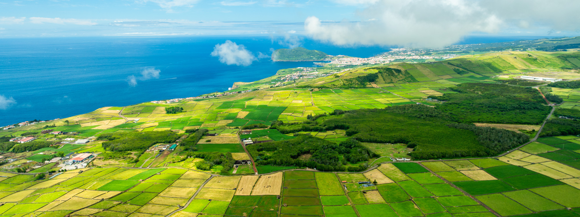 Panoramic aerial view across the green patchwork fields of Terceira Island, with rural settlements, forested areas and the Atlantic coastline stretching into the distance.