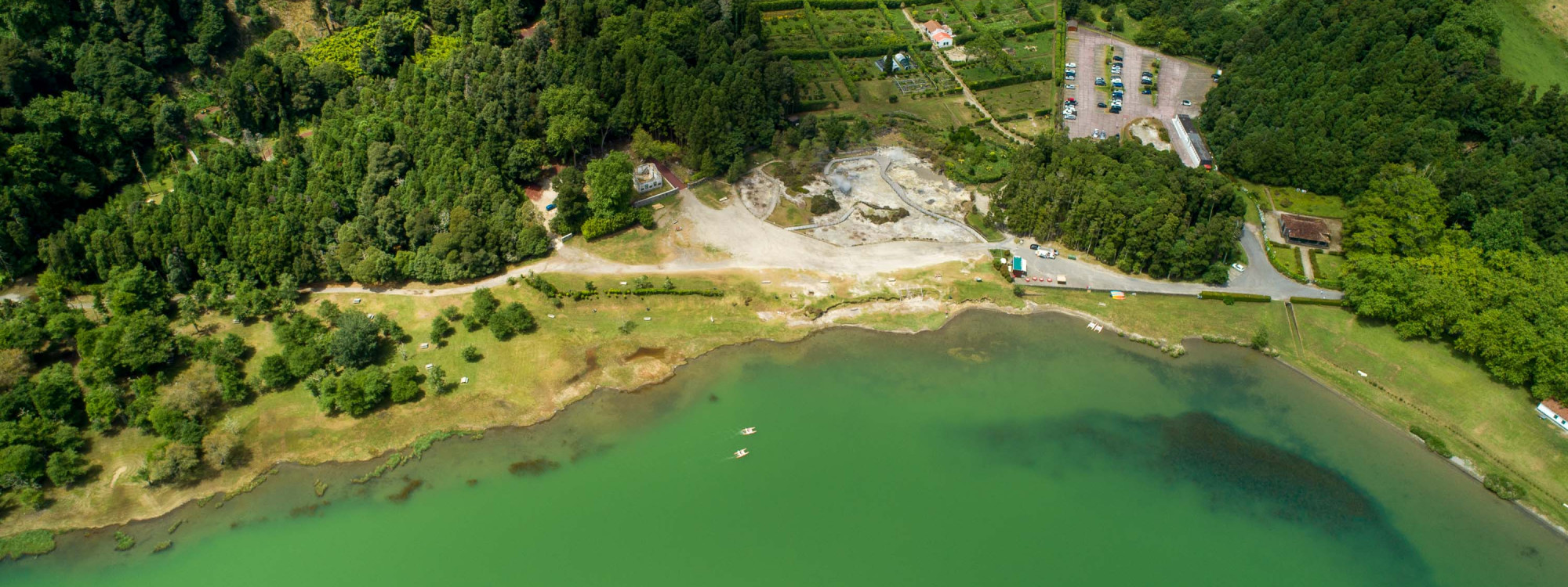 A wide aerial view shows the fumaroles and geothermal grounds along the shore of Lagoa das Furnas in São Miguel Island, Portugal. Green lake water, woodland and walking areas surround the volcanic site.
